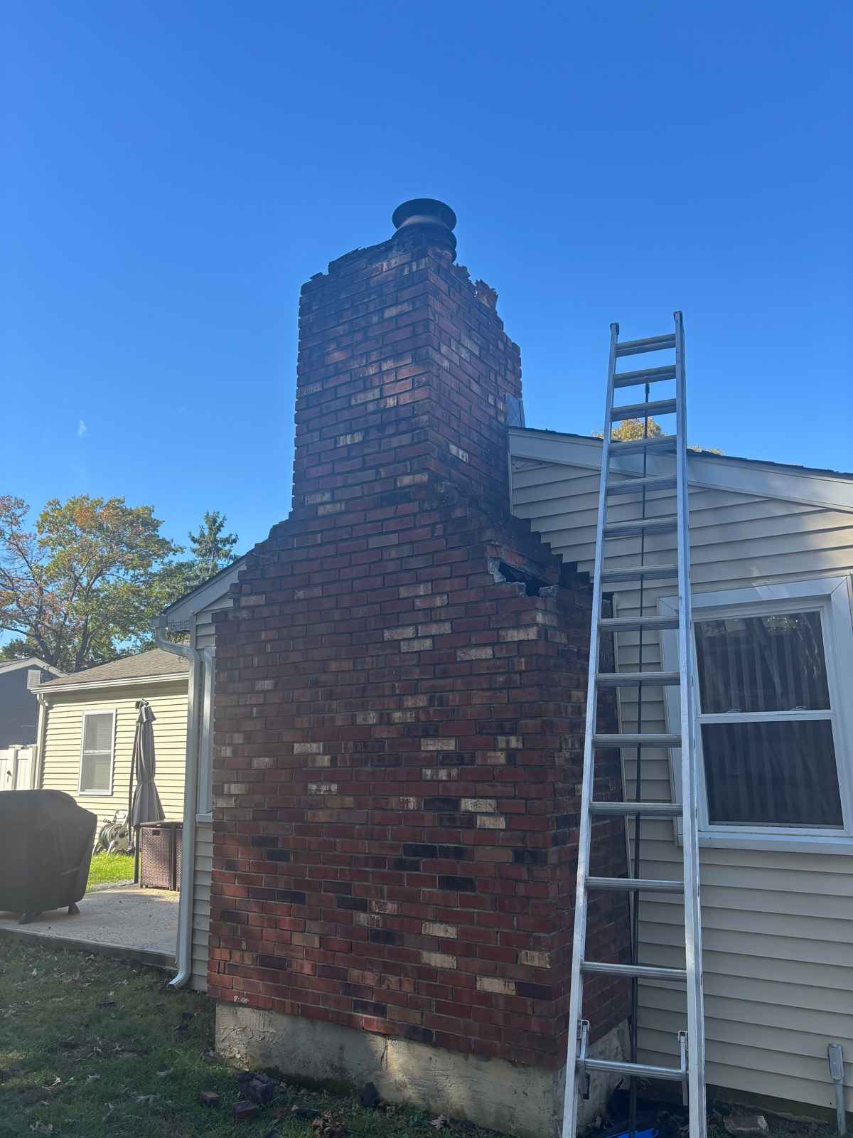 Tall exterior brick chimney on the gable wall of a house, with a ladder set against the wall, before removal